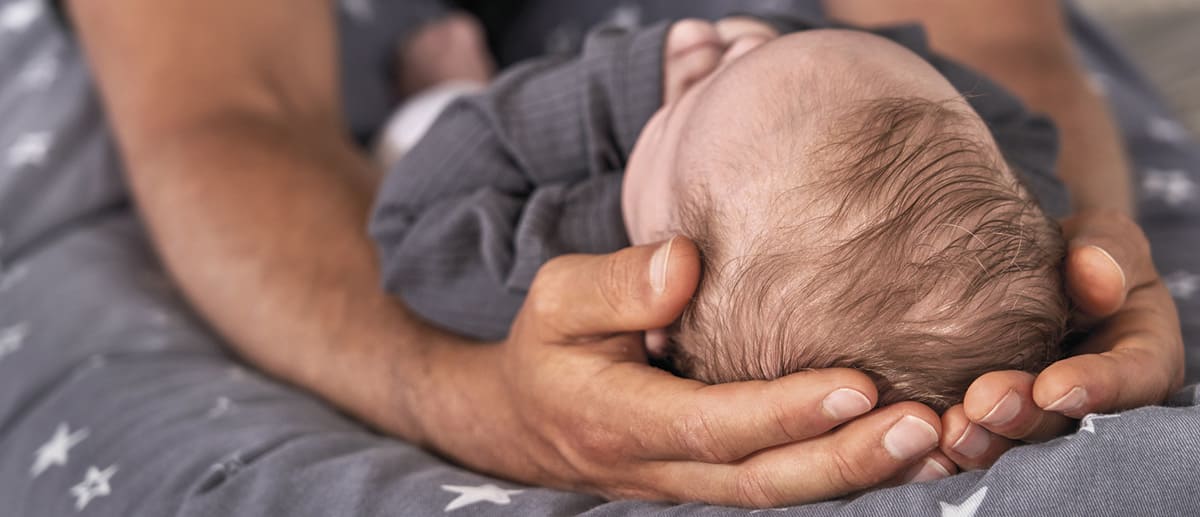 Ein Baby liegt in den Händen eines Erwachsenen und schläft auf einem grauen Kissen mit weißen Sternen. Die Umgebung wirkt beruhigend und schützend.