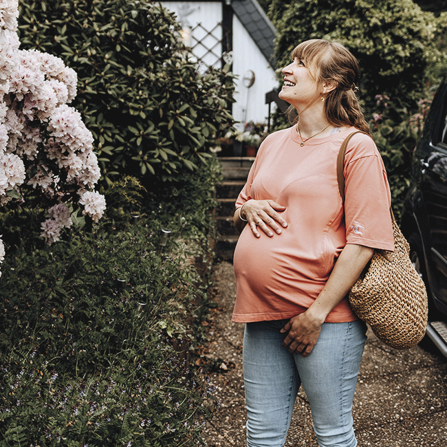 Eine schwangere Frau steht lächelnd, die Hand auf ihrem Bauch, auf einem Gartenweg. Sie trägt eine orangefarbene Bluse, blaue Jeans und eine Strohtasche, umgeben von blühenden Sträuchern.