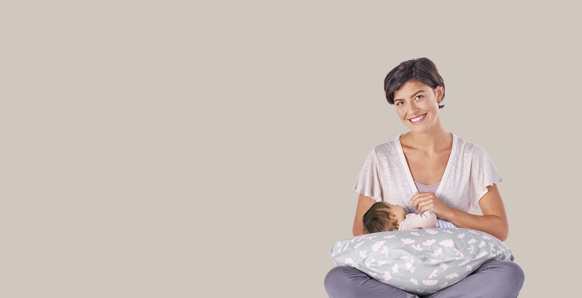 Eine Frau hält ein Baby, das auf einem Theraline-Kissen liegt. Sie sitzt lächelnd vor einem beigen Hintergrund, während das Baby mit dem Gesicht nach unten ruht.